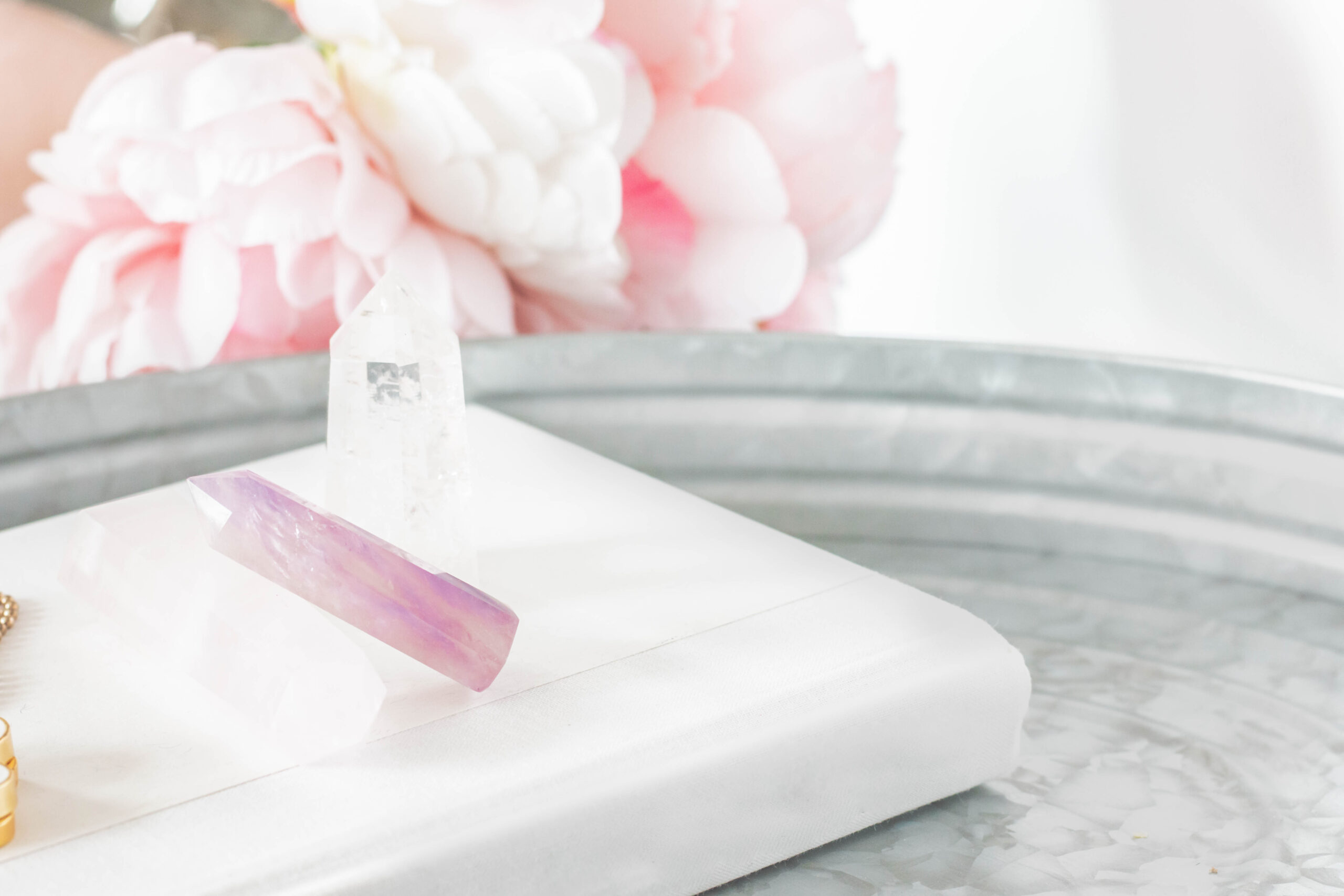 Crystals on a white surface with pink flowers blurred in the background. Soft pastel tones create a serene and calming atmosphere.