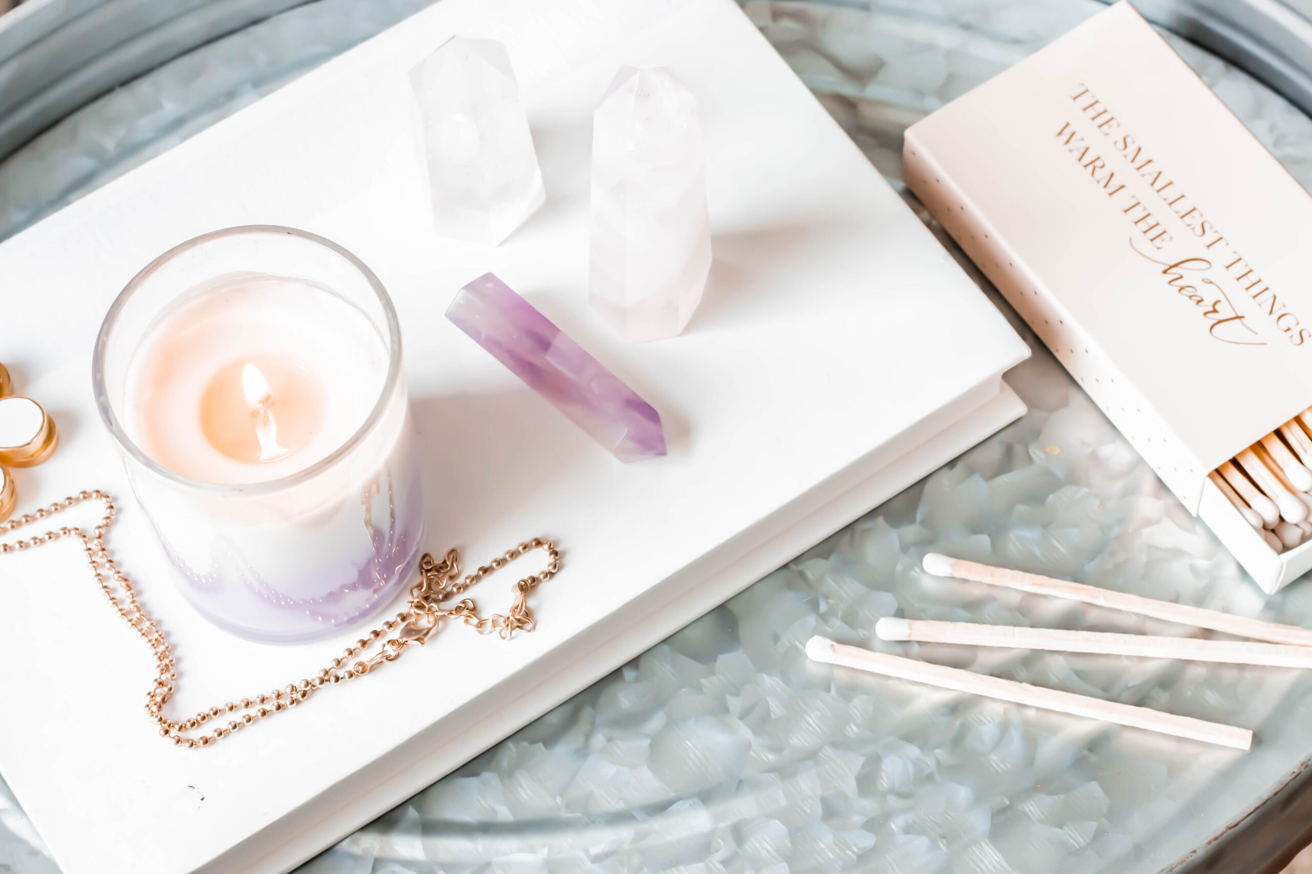 Cozy setup with a lit candle, crystals, a gold chain, and matches on a white book. Text reads "The smallest things warm the heart."