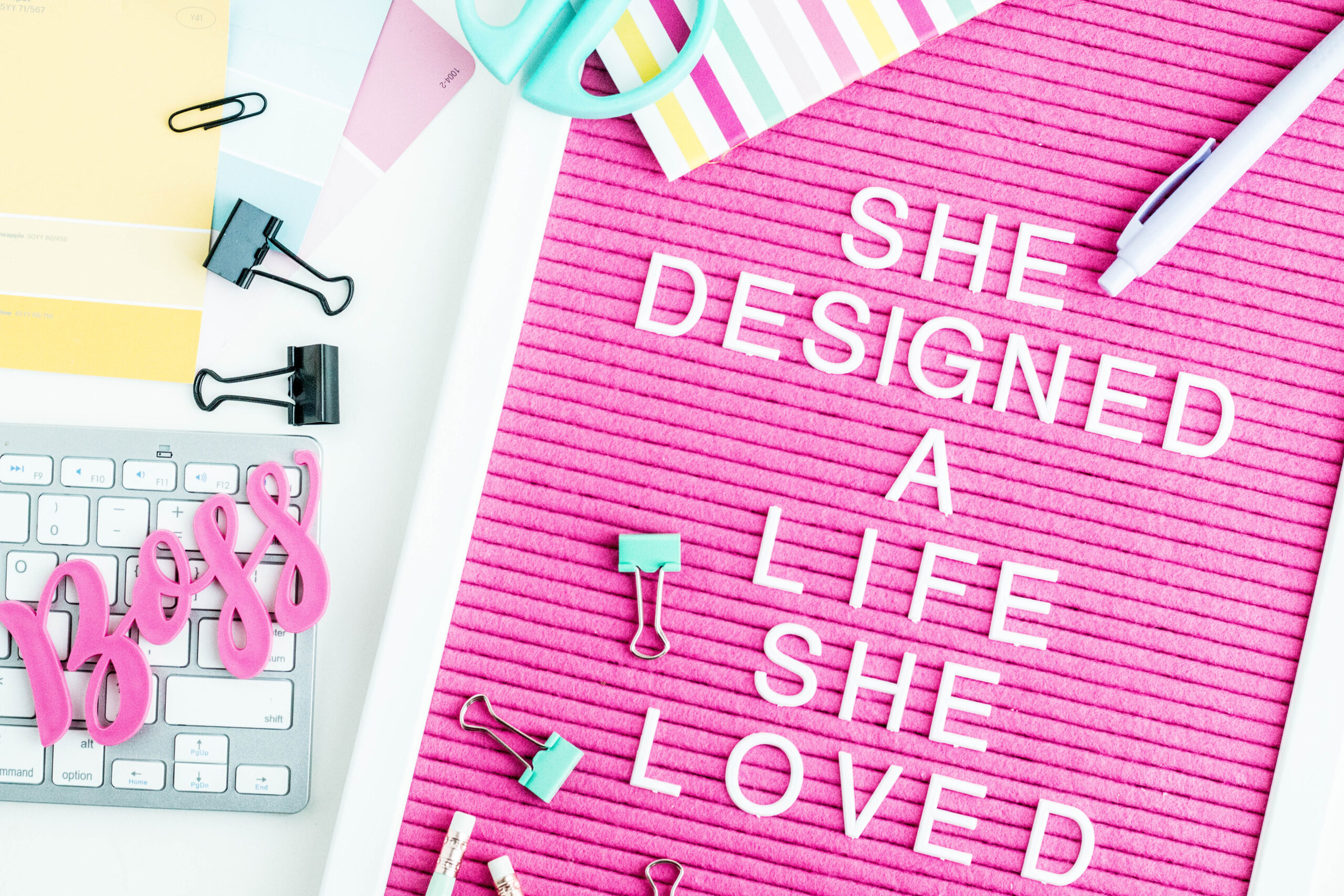 Pink letter board reads "SHE DESIGNED A LIFE SHE LOVED." Surrounding are colorful papers, a keyboard, pens, clips, and the word "boss."