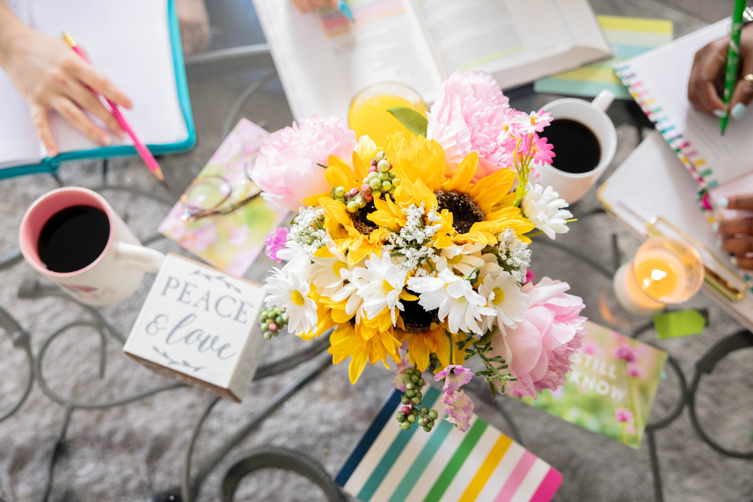Flowers on a table with books, coffee mugs, and a candle. Text reads "Peace & Love." Hands writing, colorful and inviting setting.