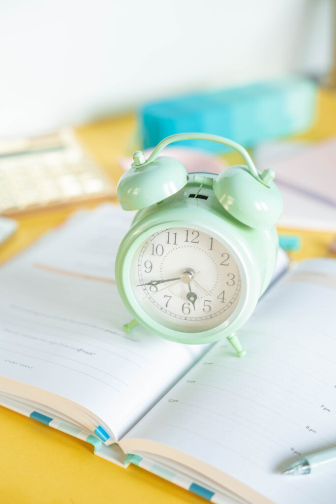 Mint-green alarm clock on an open planner, colorful blurred office supplies in the background, creating a light, organized mood.